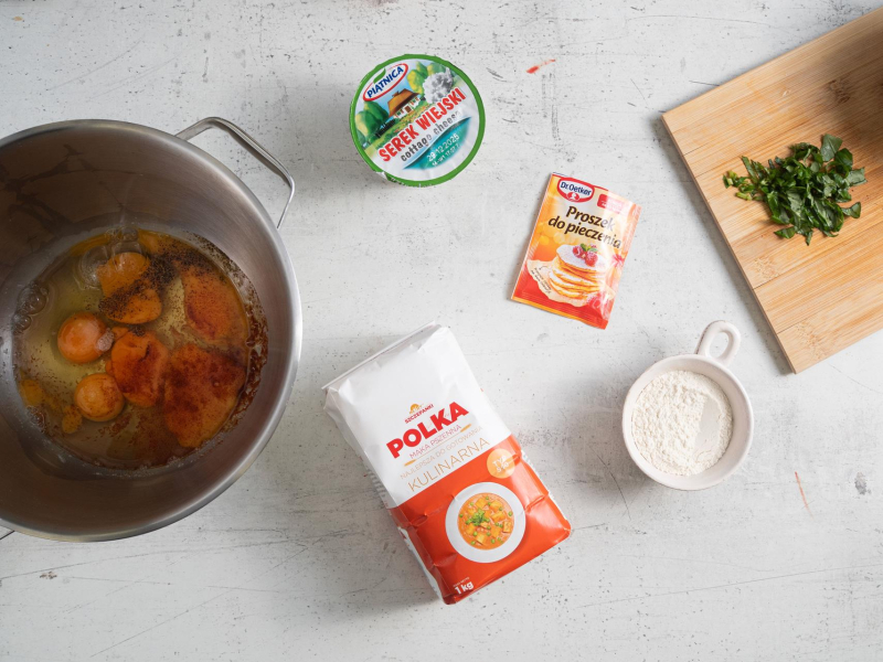 Na blacie kuchennym jajka, mąka, serek wiejski, proszek do pieczenia oraz posiekana bazylia.