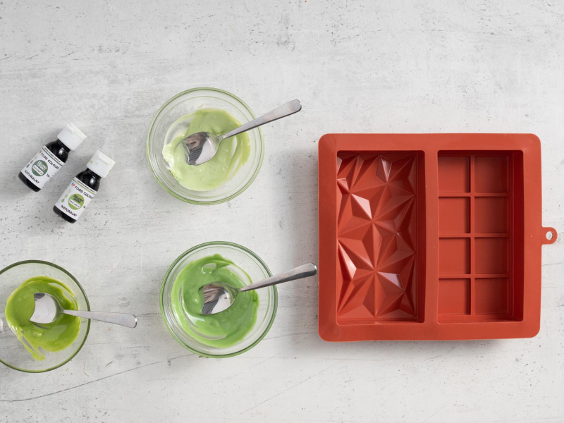 Trzy miseczki z białą czekoladą o różnym natężeniu zielonego. Obok foremka silikonowa.
