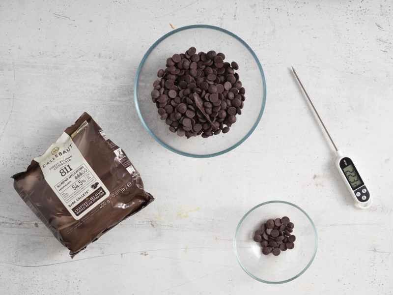 Na blacie kuchennym czekolada ciemna w kaletkach oraz termometr cukierniczy.