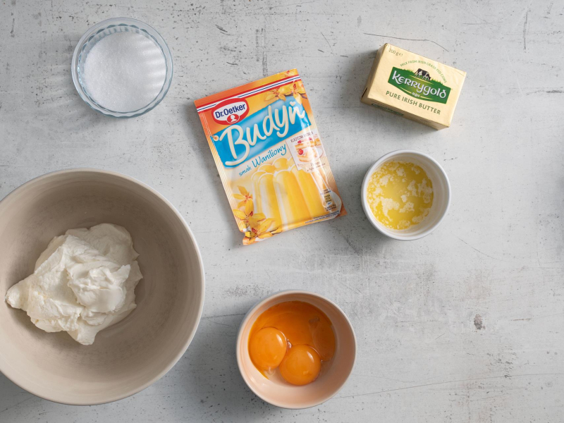 W misce skyr. Obok w miseczce cukier, 偶贸艂tka, budy艅 waniliowy oraz rozpuszczone mas艂o.