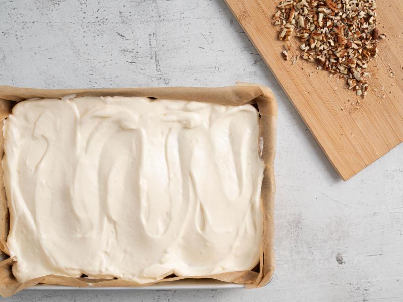 Na blacie ciasto marchewkowe.  Obok na drewnianej desce pokrojone orzechy,