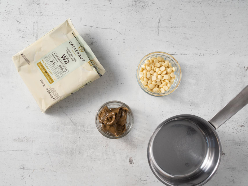 Na blacie kuchennym opakowanie białej czekolady oraz pasta pistacjowa i garnek.