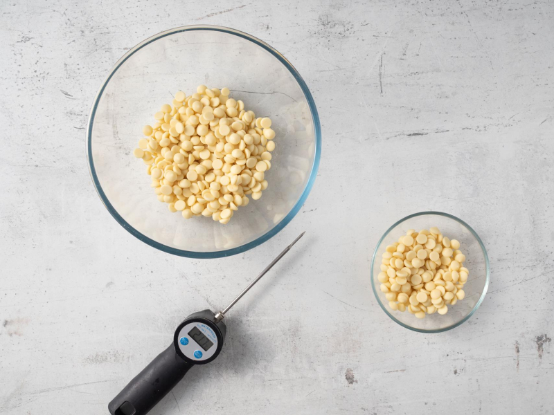 Dwie miseczki z białą czekoladą oraz termometr cukierniczy.