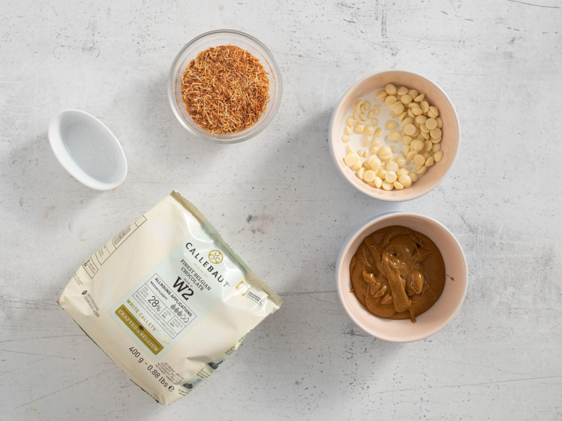 Na blacie kuchennym opakowanie białej czekolady. W miseczkach pasta pistacjowa, czekolada ze śmietanką, prażynka z ciasta kataifi oraz sól.