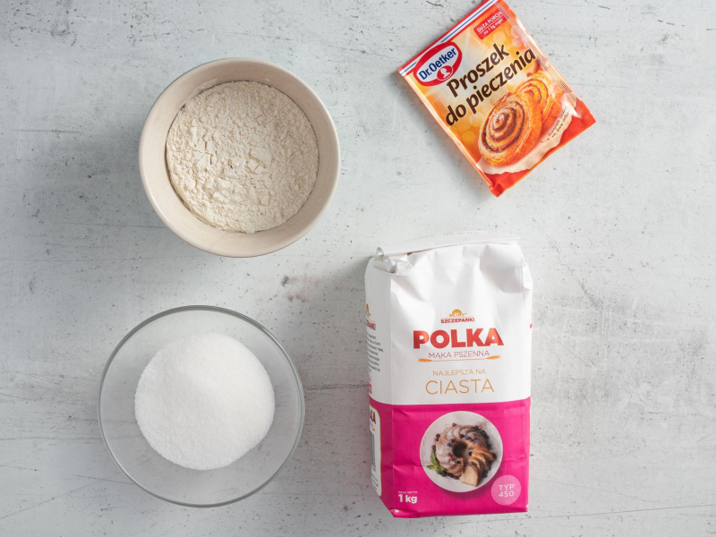 Na blacie kuchennym mąka pszenna, opakowanie proszku do pieczenia oraz cukier w miseczce.