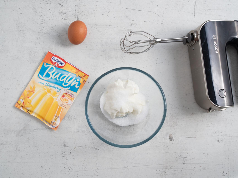 Na blacie kuchennym w misce skyr ze słodzikiem. Obok jajko, opakowanie budyniu i smaku waniliowym oraz mikser ręczny.