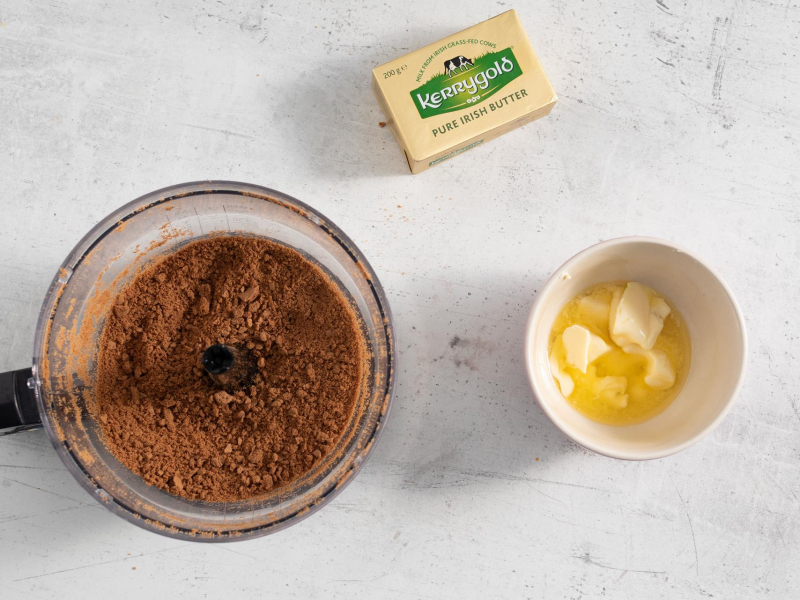 Na blacie kuchennym pokruszone ciasteczka w malakserze. Obok opakowanie masła oraz miękkie masło w miseczce.