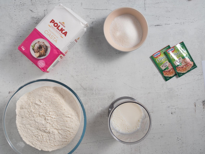 Na blacie kuchennym w miseczkach mąka pszenna, cukier, mleko oraz drożdże.