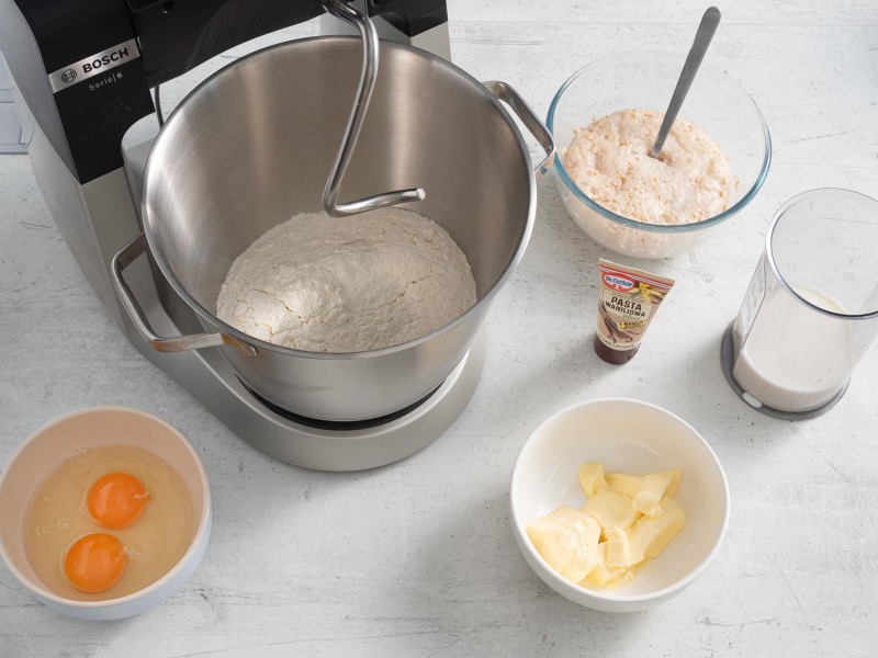 W misie wmieszane suche składniki. Obok opakowanie pasty waniliowej, jajka w misie, masło w misie, wyrośnięte drożdże oraz mleko.