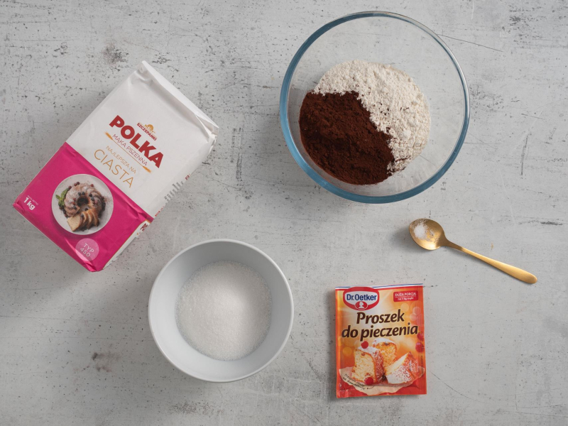 Na blacie kuchennym opakowanie mąki pszennej, proszek do pieczenia, łyżeczka soli. Obok w misce mąka z kakao oraz miseczka z cukrem.