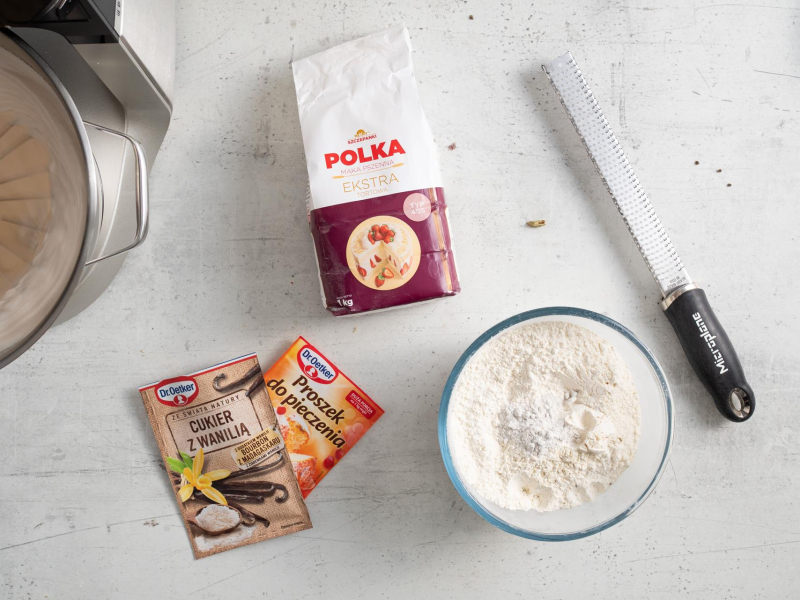 Na blacie kuchennym w misce mąka pszenna . Obok opakowanie mąki, cukru z wanilią oraz proszku do pieczenia.
