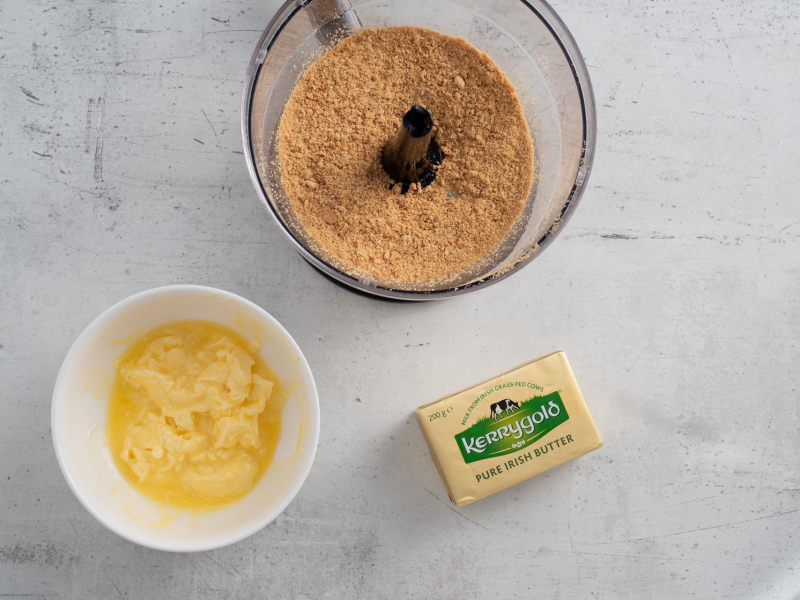 Zblendowane ciasteczka w pojemniku blendera ręcznego. Obok rozpuszczone masło.