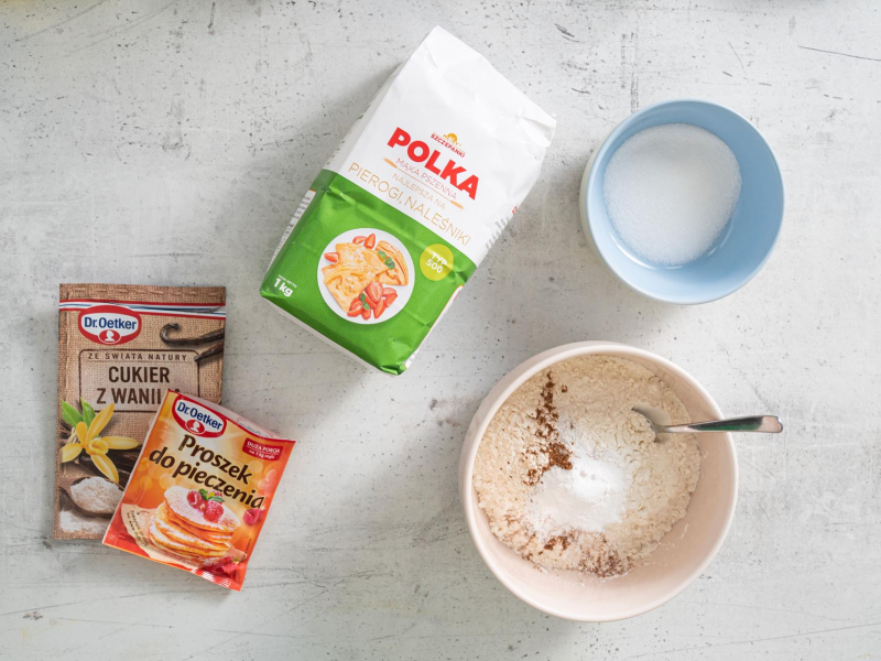 Na blacie kuchennym opakowanie mąki, proszku do pieczenia i cukru z wanilią. W miseczce cukier.