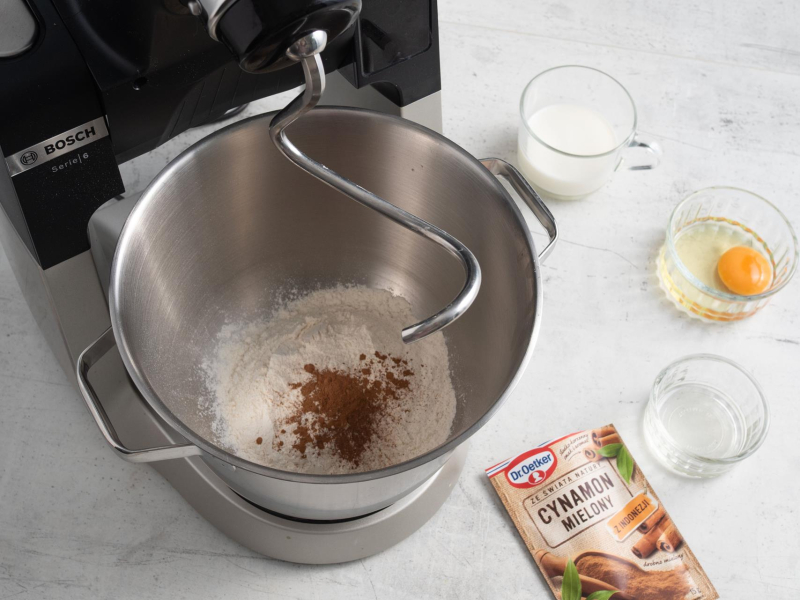 W misie robote kuchennego suche składniki do racuchów z airfryer. Na blacie kuchennym opakowanie cynamonu, jajko, mleko oraz olej w miseczkach.