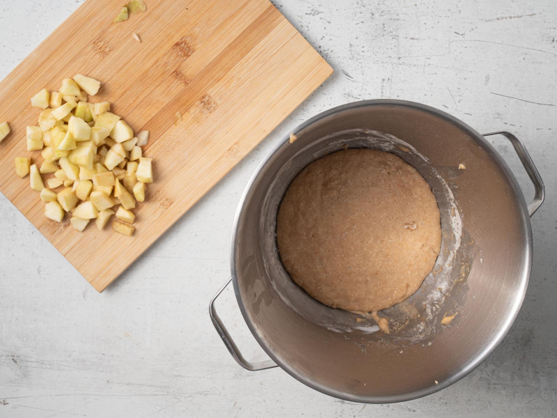 Na drewnianej desce pokrojone na małe kawałki jabłka. Obok wyrośnięte ciasto.