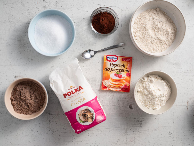 Na blacie kuchennym suche składniki do przygotowania proteinowego ciasta czekoladowego - mąka pszenna, mąka owsiana, proszek do pieczenia, sól, kakao, odżywka białkowa i erytrytol.