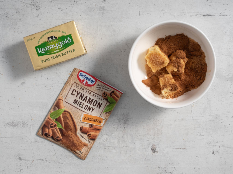 Na blacie kuchennym opakowanie masła, cynamon w opakowaniu, a w miseczce masło wymieszane z cukrem i cynamonem.