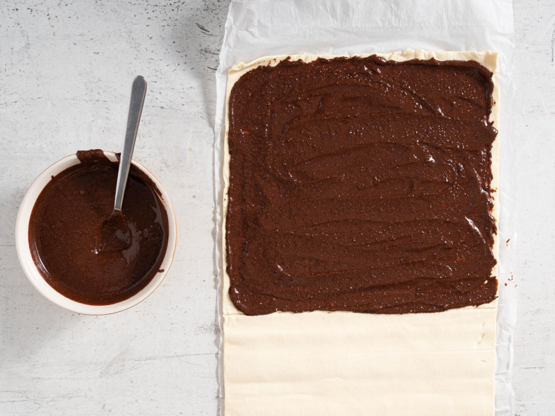 Na blacie kuchennym rozłożone ciasto francuskie posmarowane do połową kremem czekoladowym. W misce krem czekoladowy.