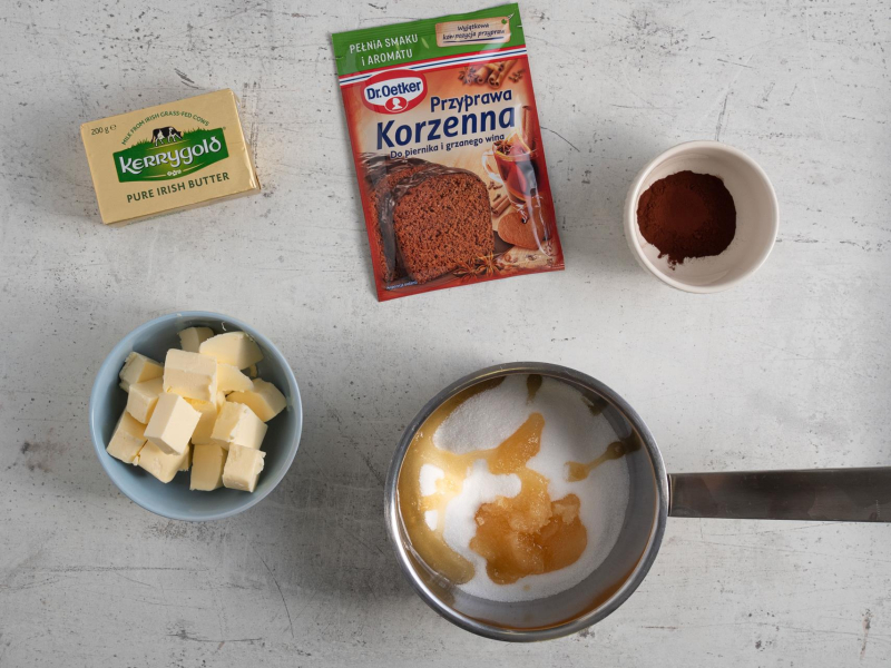 Pokrojone masło w garnku. Obok przyprawa korzenna, miód, cukier oraz kakao.