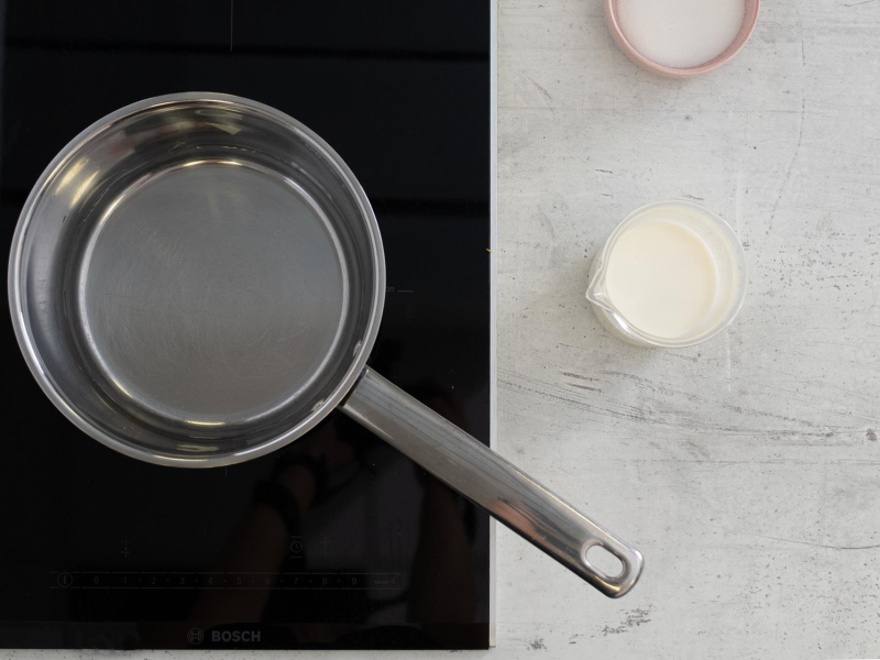 Garnek na palniku. Obok mleko w szklance oraz cukier.