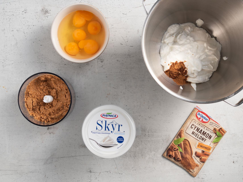 W misie skyr oraz cynamon. Obok rozdrobione ciasteczka, jajka, cynamon i opakowanie skyru.
