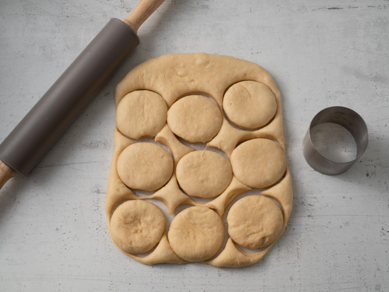 ciasto drożdżowe rozwałkowane na blacie z wyciętymi pączkami obok wałek