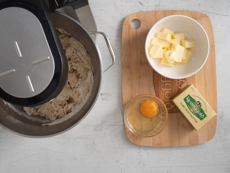 W misie miksera zagniatanie ciasta drożdżowego. Obok na desce miękkie masło oraz jajko.