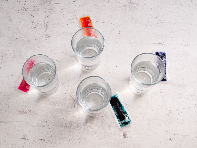4 szklanki z gorącą wodą, obok saszetki z barwnikami do jajek