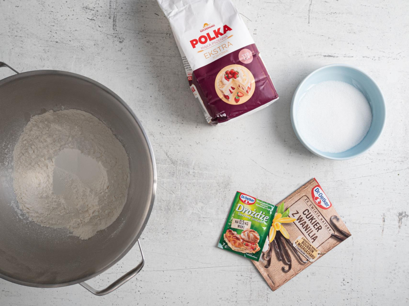 Koło robota kuchennego opakowanie masła, rozpuszczone masło w misie oraz zagniecione ciasto w mikserze.