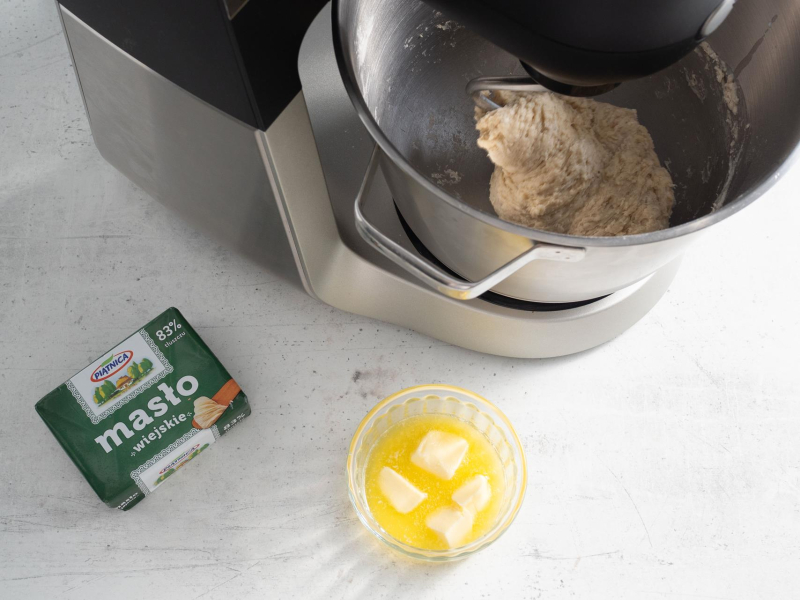 Koło robota kuchennego opakowanie masła, rozpuszczone masło w misie oraz zagniecione ciasto w mikserze.