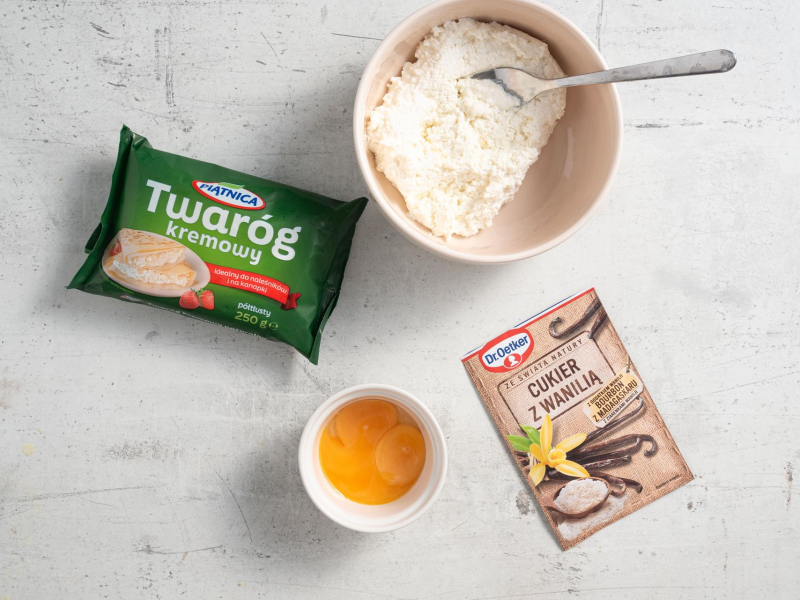 Na blacie kuchennym twaróg, żółtka w miseczce, cukier z wanilią oraz twaróg w misce.