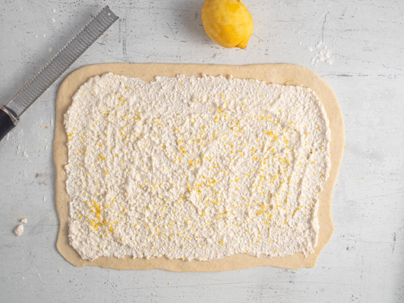 Rozwałkowane ciasto drożdżowe posmarowane twarogiem ze skórką cytrynową.