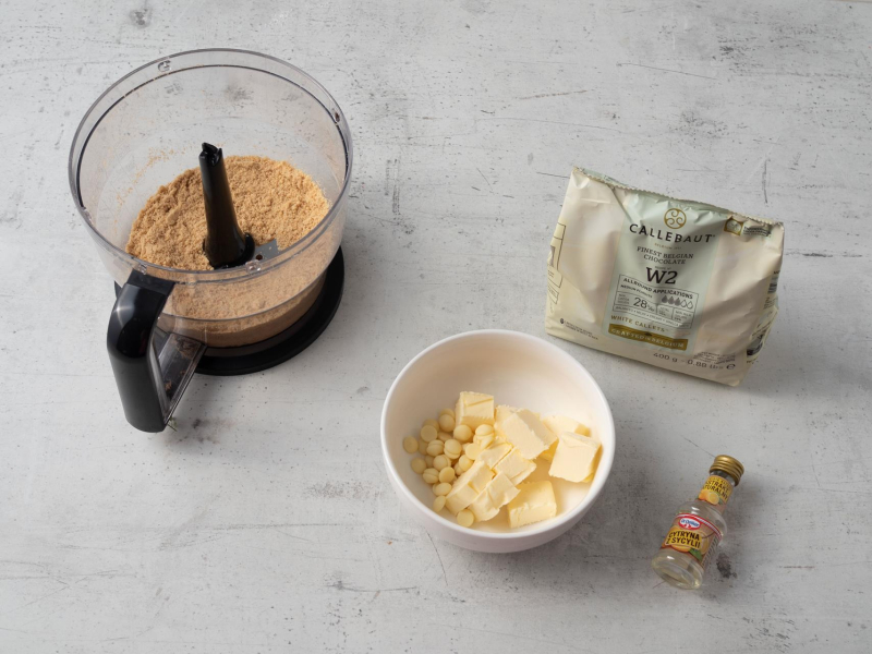 w malakserze pokruszone ciasteczka obok miseczka  z masłem, białą czekoladą i ekstraktem