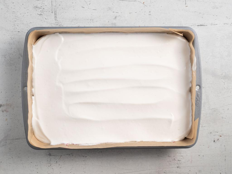 W prostokątnej blasze rozsmarowany krem z serka wiejskiego na czekoladowym fit cieście.