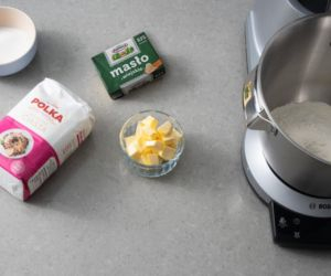 Na blacie kuchennym mąka, kostka masła oraz masło w miseczce. Obok robot kuchenny.