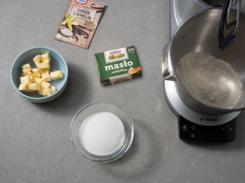 mąka i pokrojone w kostkę masło w szklanej misce, obok miseczka z cukrem i opakowanie cukru z wanilią dr. oetkera