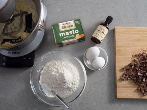 na blacie składniki do przygotowania ciasteczek piegusków: posiekana czekolada mleczna na drewnianej desce, dwa jajka w miseczce, mąka z proszkiem do pieczenia w szklanej misce, cukier w kubku, opakowanie ekstraktu waniliowego taylor and colledge oraz utarte masło w misie robota bosch