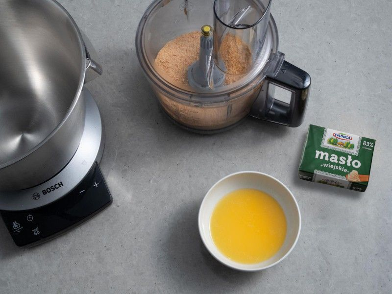 Blender ręczny firmy Bosch leżący na blacie kuchennym, w nim herbatniki. Obok miseczka z roztopionym masłem i kostka
