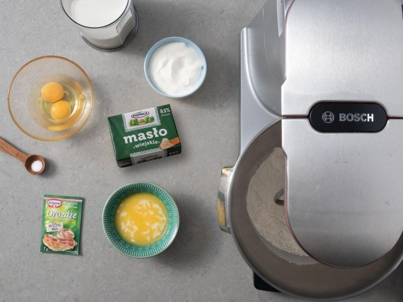 Na blacie kuchennym kostka masła, obok rozpuszczone masło w miseczce, 2 jajka w miseczce, kwaśna śmietana oraz mleko i suszone drożdże