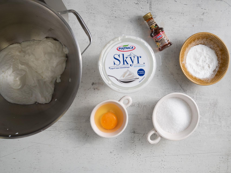 Na blacie kuchennym składniki na masę serową: skyr, jajko, ksylitol, ekstrakt waniliowy i mąka ziemniaczana.