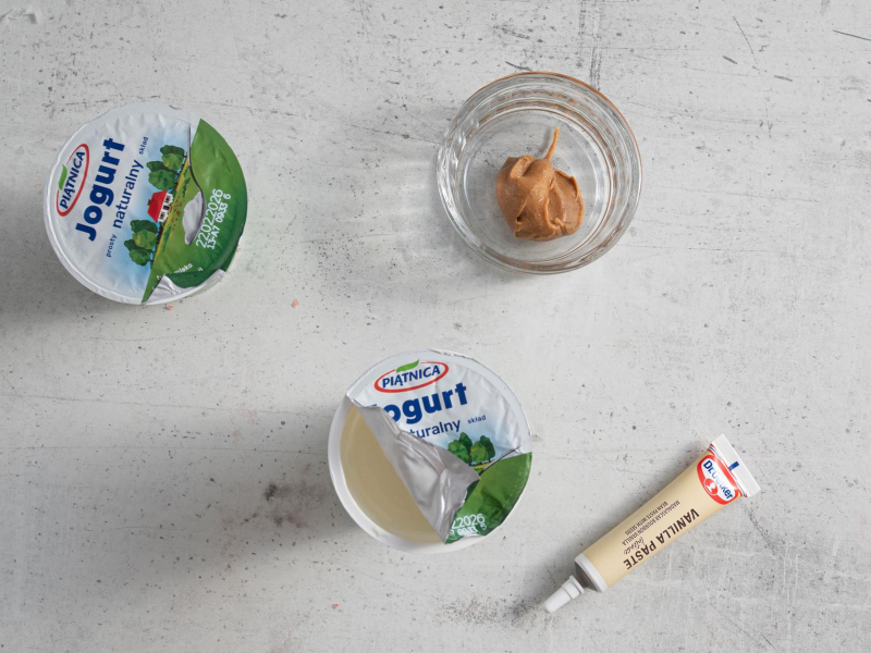 Na blacie kuchennym opakowania jogurtu naturalnego oraz masło orzechowe w miseczce i pasta z wanilii.