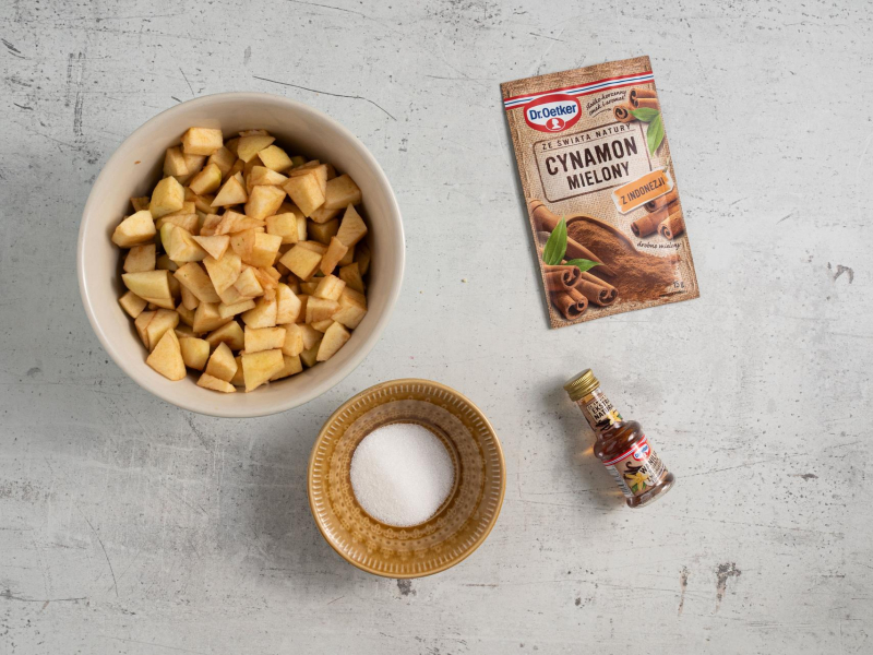 W misce pokrojone jabłka. Obok cukier w miseczce, cynamon mielony i ekstrakt waniliowy.